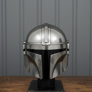 Puede incluir: Un casco plateado de Mandaloriano, exhibido sobre un soporte negro. El casco tiene una parte superior redondeada con crestas verticales y detalles faciales angulares. El fondo es una pared gris y una mesa de madera.