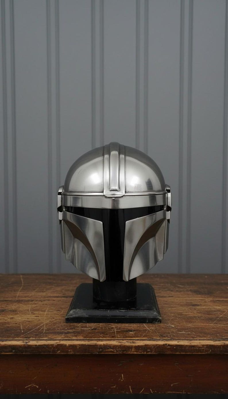 Puede incluir: Un casco plateado de Mandaloriano, exhibido sobre un soporte negro. El casco tiene una parte superior redondeada con crestas verticales y detalles faciales angulares. El fondo es una pared gris y una mesa de madera.