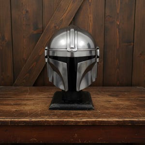 Puede incluir: Un casco plateado de Mandaloriano, exhibido sobre un soporte negro. El casco tiene un acabado pulido con paneles detallados y una visera distintiva. El casco se encuentra sobre un fondo de madera.