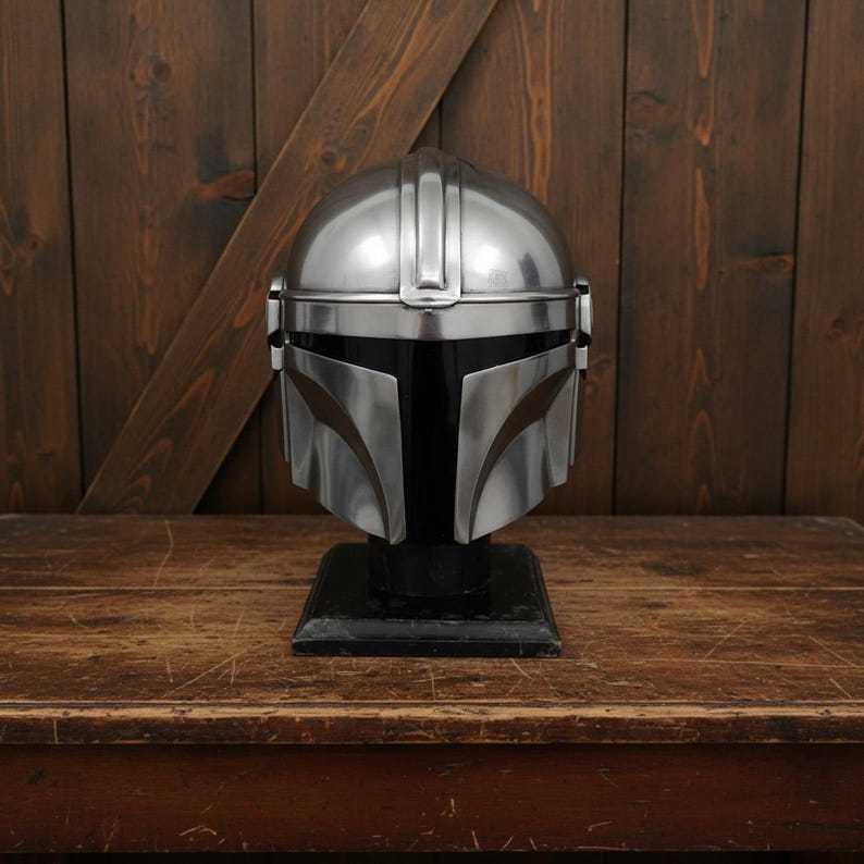 Puede incluir: Un casco plateado de Mandaloriano, exhibido sobre un soporte negro. El casco tiene un acabado pulido con paneles detallados y una visera distintiva. El casco se encuentra sobre un fondo de madera.