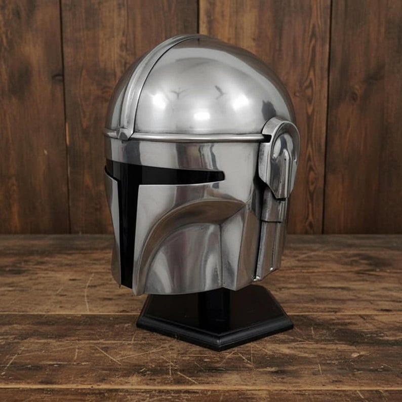 Puede incluir: Un casco plateado pulido con visera negra y orejeras, exhibido sobre un soporte negro. El casco tiene una parte superior redondeada y rasgos angulares, que reflejan la luz. El dise&ntilde;o recuerda a un personaje espacial.