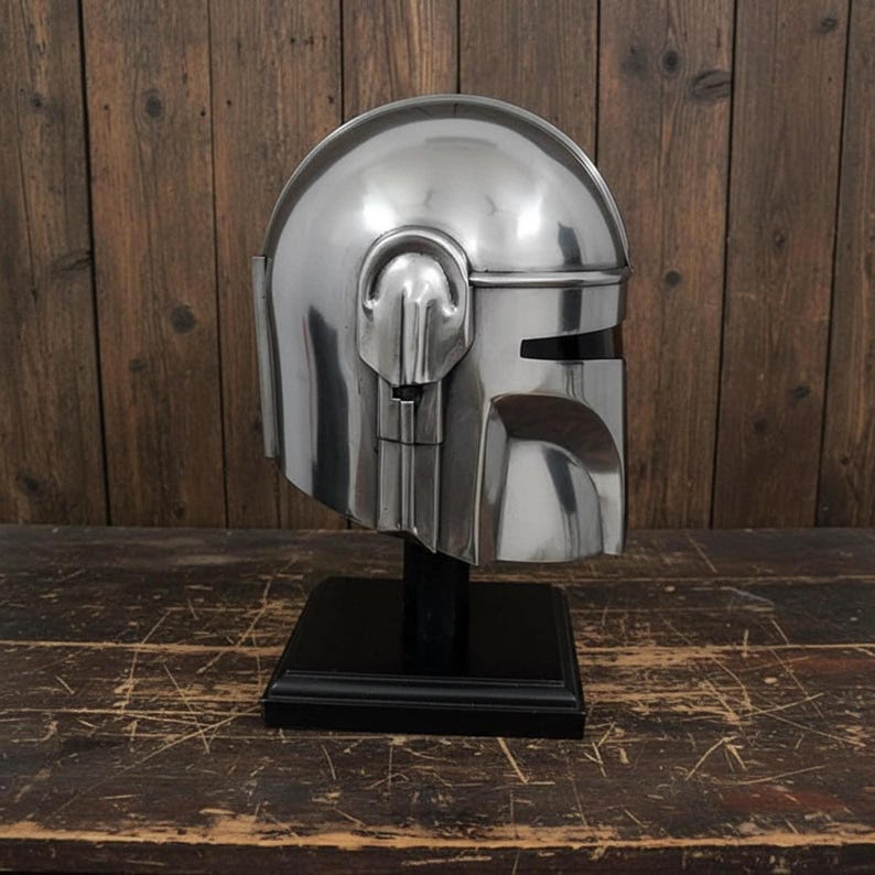 Puede incluir: Un casco plateado con una visera y orejeras distintivas, exhibido sobre un soporte negro. El casco tiene una superficie lisa y reflectante, sobre un fondo de madera. El dise&ntilde;o sugiere un tema futurista o de ciencia ficci&oacute;n.