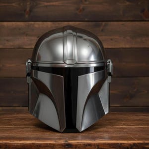 Puede incluir: Un casco plateado de Mandaloriano con visera negra, sobre una superficie de madera y fondo oscuro. El casco, de dise&ntilde;o angular, presenta un acabado pulido y un aspecto elegante.