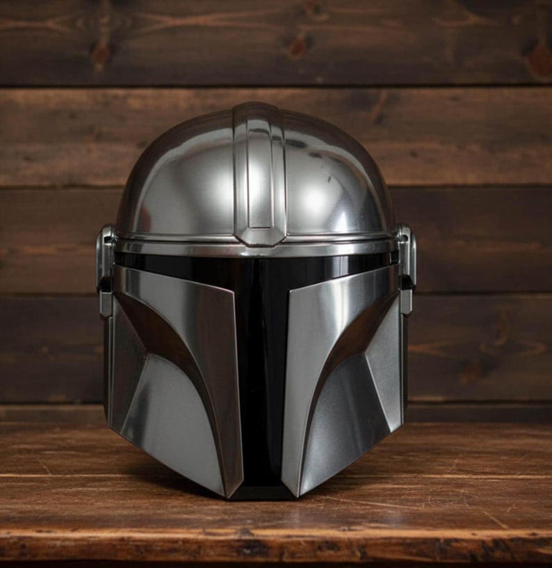 Puede incluir: Un casco plateado de Mandaloriano con visera negra, sobre una superficie de madera y fondo oscuro. El casco, de dise&ntilde;o angular, presenta un acabado pulido y un aspecto elegante.