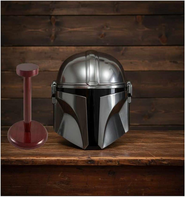 Puede incluir: Un casco plateado mandaloriano con una visera negra sobre una superficie de madera. Un soporte de casco granate a la izquierda. El casco es una r&eacute;plica detallada de la franquicia Star Wars.