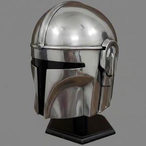 Puede incluir: Un casco plateado pulido con una visera negra, exhibido sobre un soporte negro. El casco tiene una parte superior redondeada con una cresta distintiva y detalles angulares alrededor de la cara y los lados. El dise&ntilde;o sugiere un tema futurista o de ciencia ficci&oacute;n.