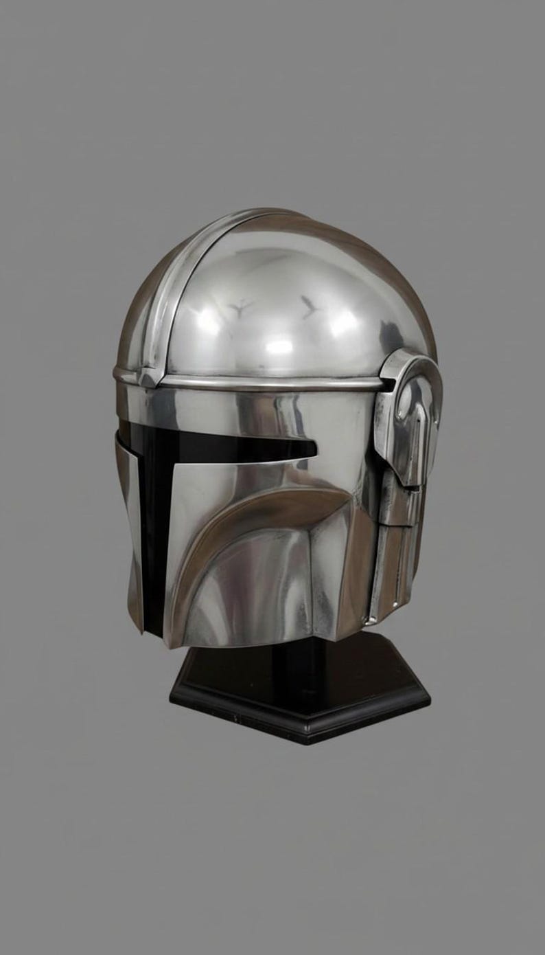 Puede incluir: Un casco plateado pulido con una visera negra, exhibido sobre un soporte negro. El casco tiene una parte superior redondeada con una cresta distintiva y detalles angulares alrededor de la cara y los lados. El dise&ntilde;o sugiere un tema futurista o de ciencia ficci&oacute;n.