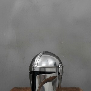 Puede incluir: Un brillante casco plateado mandaloriano con una distintiva visera en forma de T, apoyado sobre una mesa de madera r&uacute;stica. La superficie pulida del casco refleja la luz, resaltando su dise&ntilde;o detallado. El fondo es una pared gris lisa.