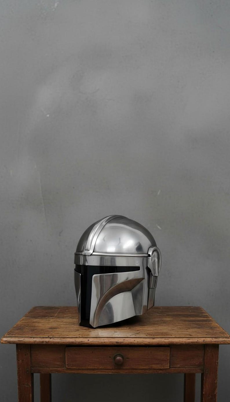 Puede incluir: Un brillante casco plateado mandaloriano con una distintiva visera en forma de T, apoyado sobre una mesa de madera r&uacute;stica. La superficie pulida del casco refleja la luz, resaltando su dise&ntilde;o detallado. El fondo es una pared gris lisa.