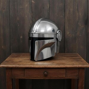 Puede incluir: Un casco plateado pulido con una distintiva visera en forma de T, apoyado sobre una mesa de madera. El dise&ntilde;o del casco presenta formas angulares y una visera curva, sugiriendo un tema futurista o de ciencia ficci&oacute;n. El fondo es una pared de madera oscura.