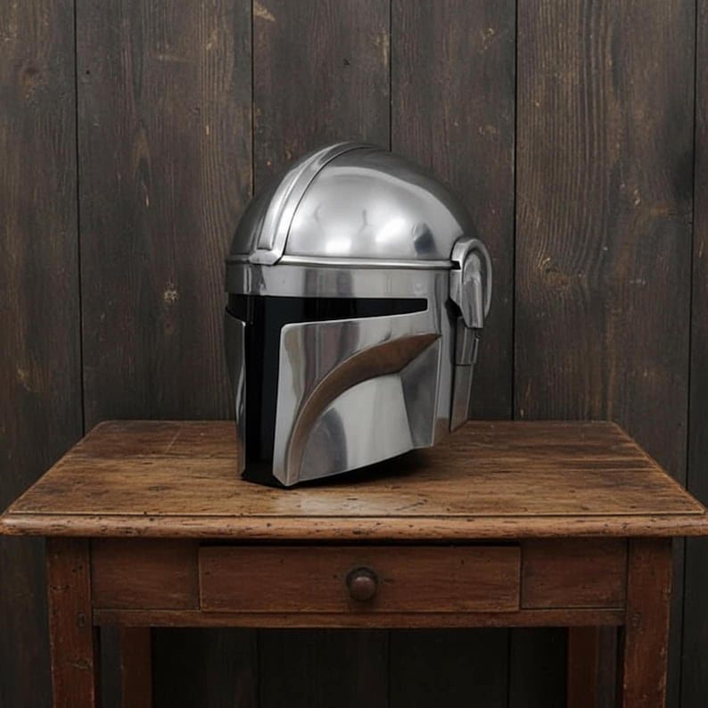 Puede incluir: Un casco plateado pulido con una distintiva visera en forma de T, apoyado sobre una mesa de madera. El dise&ntilde;o del casco presenta formas angulares y una visera curva, sugiriendo un tema futurista o de ciencia ficci&oacute;n. El fondo es una pared de madera oscura.