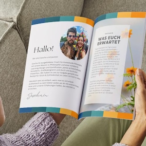 以下が含まれることがあります： 「Hallo!」と「Was Euch Erwartet」というドイツ語のテキストが書かれた開いた本を人が持っています。本にはカップルの写真とテキストが掲載されています。右ページにはオレンジ色の花が見えます。本にはカラフルなボーダーがあります。
