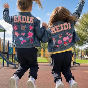 Könnte beinhalten: Zwei blaue Jeansjacken mit den personalisierten Namen SADIE und HEIDI in großen rosa Buchstaben auf dem Rücken. Jede Jacke ist mit verschiedenen bunten Stickaufnähern verziert, darunter Herzen, eine Meerjungfrauenflosse, ein Pilz und ein Haus.
