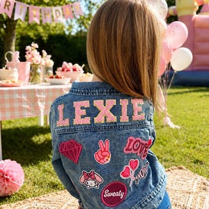 Könnte beinhalten: Eine blaue Jeansjacke mit dem Namen LEXIE in großen, rosa glitzernden Buchstaben auf dem Rücken. Die Jacke ist mit verschiedenen gestickten Aufnähern verziert, darunter ein rosa Diamant, ein Friedenszeichen, das Wort LOVE, Herzen, eine Comicfigur und das Wort Sweaty.