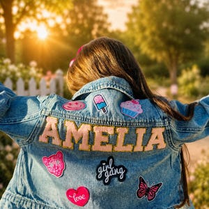 May include: A light blue denim jacket with the name "AMELIA" in pink letters. The jacket is decorated with patches including a pink heart with "Love", a butterfly, and a "girl gang" patch. The jacket is worn outdoors.