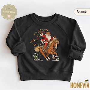 May include: A black long-sleeve sweatshirt featuring a graphic print of Santa Claus wearing a cowboy hat and red suit, riding a brown horse while holding a string of colorful Christmas lights. Two small green cacti are visible at the bottom.