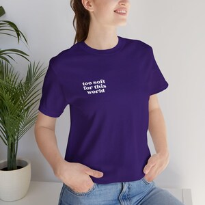 Könnte beinhalten: Ein dunkelviolettes Kurzarm-T-Shirt mit dem weißen Schriftzug "too soft for this world" auf der linken Brustseite. Das Oberteil verfügt über einen Rundhalsausschnitt und eine klassische Passform für einen schlichten und klaren Look.