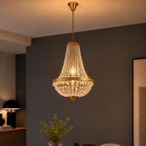 Puede incluir: Una lámpara de araña estilo imperio en tono dorado con cuentas de cristal transparente en cascada y un marco de metal decorativo. La pieza está suspendida por una cadena dorada a juego y emite una luz cálida desde sus bombillas internas.