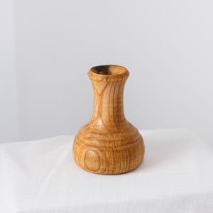 May include: A small, hand-turned wooden vase with a natural light brown finish, featuring a wide, rounded base that tapers into a slender neck and a flared rim, showcasing the distinct grain patterns and textures of the wood throughout the surface.