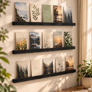May include: Three horizontal black wall-mounted floating shelves arranged in a vertical stack. Each shelf holds a collection of rectangular art prints and postcards featuring nature scenes, botanical illustrations, and text-based designs.