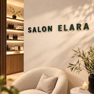 Könnte beinhalten: Eine beige Wand zeigt den dunkelgrünen Schriftzug SALON ELARA in erhabenen Buchstaben. Darunter steht ein geschwungener, strukturierter beiger Sessel mit passendem Kissen neben einem runden Marmortisch mit einer Kerze und einer Pflanze.