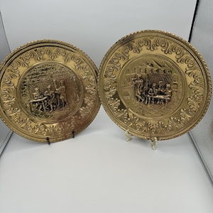May include: Two round brass decorative plates featuring embossed scenes of figures gathered around tables in an interior setting. The plates have wide, ornate borders with scrolling patterns and a polished metallic finish, displayed on small stands.