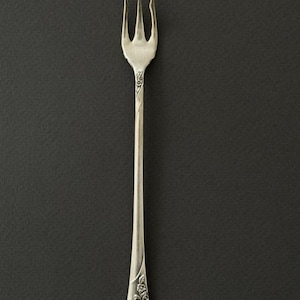 May include: A silver fork with three prongs, set against a dark background. The handle has an ornate floral design. The fork is likely made of stainless steel or a similar metal.