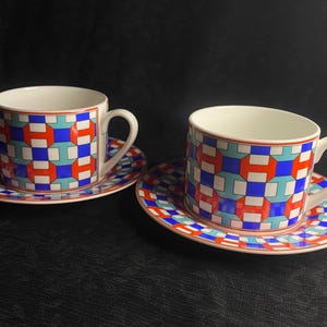Puede incluir: Dos tazas de té de cerámica con sus platos a juego que presentan un patrón geométrico repetitivo en tonos vibrantes de naranja, azul real, azul claro y blanco. Cada taza tiene un asa curva y un interior blanco liso sobre fondo oscuro.