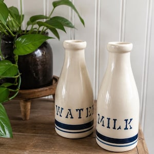 May include: Two off-white ceramic bottles with narrow necks and flared rims. One bottle features the blue text WATER and the other features the blue text MILK. Both bottles have two horizontal blue stripes near the base and a smooth finish.