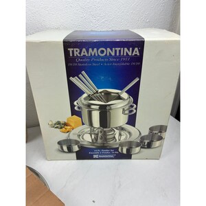 May include: A Tramontina 14-piece fondue set in a white box. The box features a graphic of a stainless steel fondue pot, forks, and small bowls. Text on the box reads Tramontina, Quality Products Since 1911, 18/10 Stainless Steel, and 14 Pc. Fondue Set.