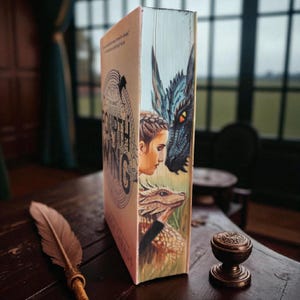 Op de afbeelding: Een hardcover boek getiteld "Fourth Wing" met een gedetailleerde illustratie van een vrouw en draken. De rug van het boek toont een drakenkop en een kleinere draak. Een veer en een inktpot staan op een houten tafel, als aanvulling op het fantasiethema van het boek.