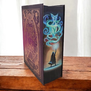 Op de afbeelding: Een decoratief boek met een paarse kaft en gouden letters met de tekst "Harry Potter I-VII". De rug toont een schilderij van een tovenaar en een gloeiend blauw wezen. Het boek wordt gepresenteerd op een houten oppervlak.