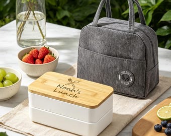 May include: A rectangular two-tier lunch box with a light wood lid engraved with the text "Noah's Lunch" and a fork and spoon icon, paired with a grey fabric insulated lunch bag featuring a circular patch with a panda graphic.