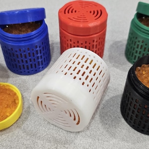 May include: A collection of six cylindrical containers in yellow, blue, red, white, green, and black. Each container features a grid-patterned body and a vented lid, filled with small, translucent orange beads visible through the openings.