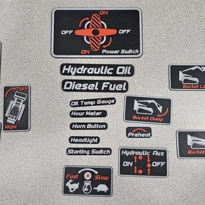 May include: A collection of black and red control panel stickers with white text and icons. The stickers include labels for hydraulic oil, diesel fuel, and various functions like bucket lift, turn, and power switch. The image is taken from above.