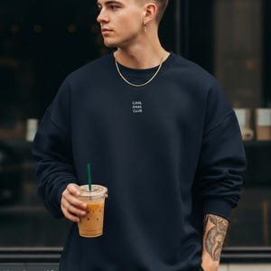 Könnte beinhalten: Ein marineblaues Sweatshirt mit Rundhalsausschnitt und dem weißen Schriftzug COOL DADS CLUB, der in einem kleinen, zentrierten Block auf der oberen Brust gedruckt ist. Das Kleidungsstück hat lange Ärmel mit gerippten Bündchen und einen gerippten Ausschnitt.