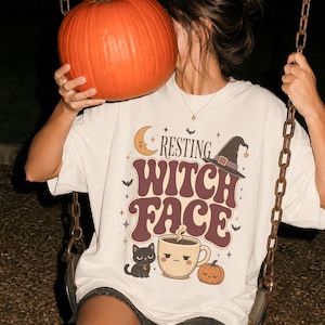 Könnte beinhalten: Ein cremefarbenes T-Shirt mit einem Grafikdruck, der den Text "RESTING WITCH FACE" in großen braunen Buchstaben zeigt. Das Design umfasst eine Mondsichel, einen Hexenhut, eine schwarze Katze, eine dampfende Kaffeetasse mit Gesicht und einen Kürbis.