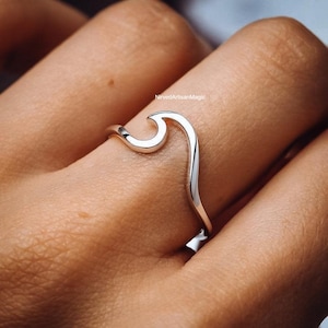 May include: A polished silver ring featuring a single, continuous wave-shaped design element that curves upwards. The metal has a smooth, reflective surface. The text NirvedArtisanMagic is visible in small, dark lettering near the wave crest.