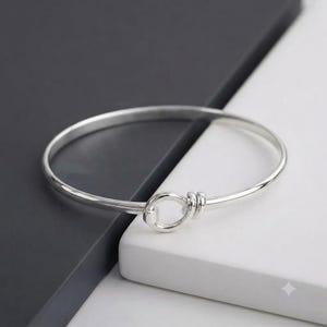 Może przedstawiać: Polerowana bransoletka typu bangle w kolorze srebrnym z mechanizmem zamykającym w formie okrągłej pętli. Bransoletka ma gładkie, odblaskowe wykończenie metaliczne oraz ciągły, okrągły kształt, który łączy się w jednym punkcie z pierścieniem.