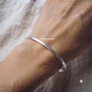 May include: A thin, polished silver bangle bracelet with a smooth, rounded surface. The metal has a reflective finish and a simple, continuous circular design. The text NirvedArtisanMagic is visible on the surface of the silver bangle.