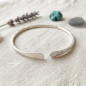 Può includere: Un bracciale a fascia in metallo color argento con una finitura martellata e testurizzata. Il bracciale presenta un design aperto con due estremità arrotondate e piatte. La superficie del metallo riflette la luce, mettendo in risalto.