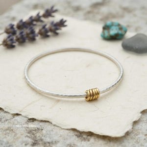 Può includere: Un bracciale rigido in argento con texture martellata caratterizzato da quattro piccoli anelli color oro che scorrono lungo la fascia. Il testo NirvedArtisanMagic è stampato nell'angolo in basso a sinistra dell'immagine, che include lavanda e pietre.