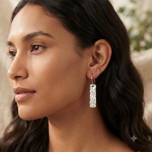 Peut inclure: Une boucle d'oreille pendante en argent dotée d'un pendentif rectangulaire à la surface martelée et texturée. Le pendentif est suspendu à un crochet simple en argent, offrant un aspect métallique propre et réfléchissant.
