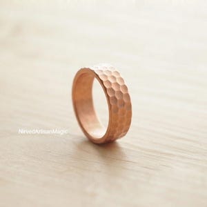 Può includere: Un anello color rame caratterizzato da una superficie martellata e testurizzata con un motivo a fossette ripetuto. La fascia ha un profilo arrotondato e una calda finitura metallica. Il testo NirvedArtisanMagic è stampato in piccole lettere.