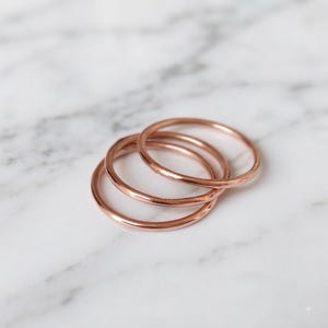 May include: A set of three thin, circular rings crafted from a polished rose gold-toned metal. The rings feature a smooth, continuous band design with a reflective surface, arranged in a slightly overlapping stack to show their rounded profile.
