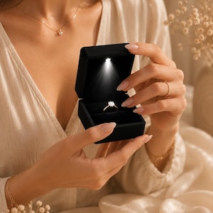 Peut inclure: Une boîte à bijoux en velours noir avec une lumière intégrée met en valeur une bague de fiançailles en diamant. La bague est sertie d'un anneau en or. La boîte est ouverte, révélant la bague. L'arrière-plan est de couleur neutre.