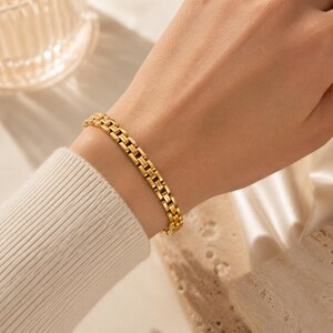 Peut inclure: Un bracelet doré avec un design à maillons rectangulaires. Le bracelet est porté au poignet, et l'image est bien éclairée, mettant en valeur les détails du bijou. Le bracelet semble être en métal et a une finition polie.
