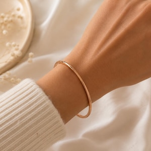 Peut inclure: Un bracelet jonc de couleur or est porté au poignet. Le bracelet est une fine bande simple. Le poignet est contre un fond blanc. La personne porte un pull crème.