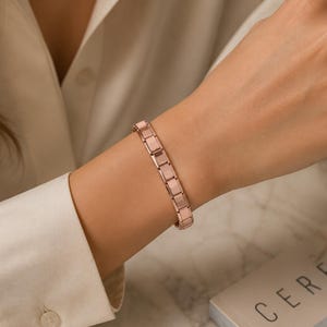 Peut inclure: Bracelet en or rose avec un design à maillons rectangulaires. Le bracelet est porté au poignet, avec une manche de chemise blanche visible. Une tasse de café et un livre sont en arrière-plan.