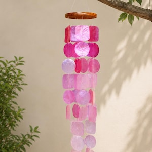 May include: A hanging wind chime featuring a circular wooden top piece with multiple strands of translucent, iridescent shell discs and squares in varying shades of pink, magenta, and light purple, suspended by thin clear nylon strings.